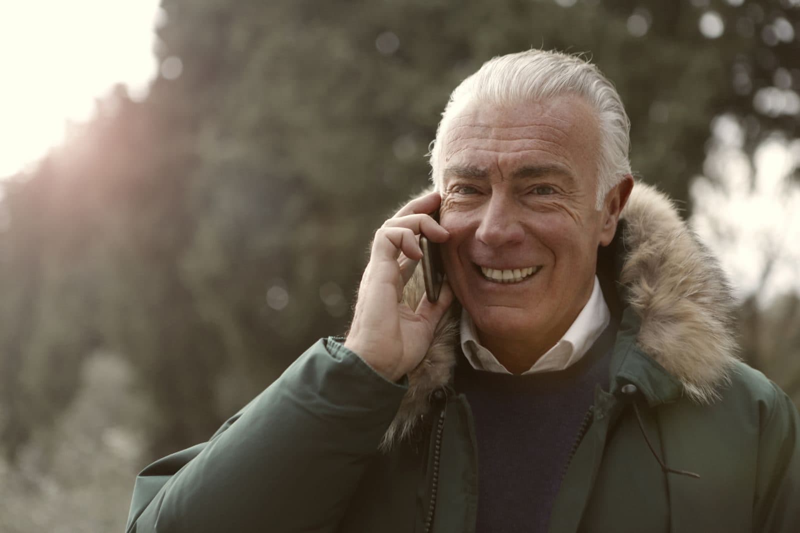 A smiling grey-haired man in a winter coat talking on a mobile phone outdoors